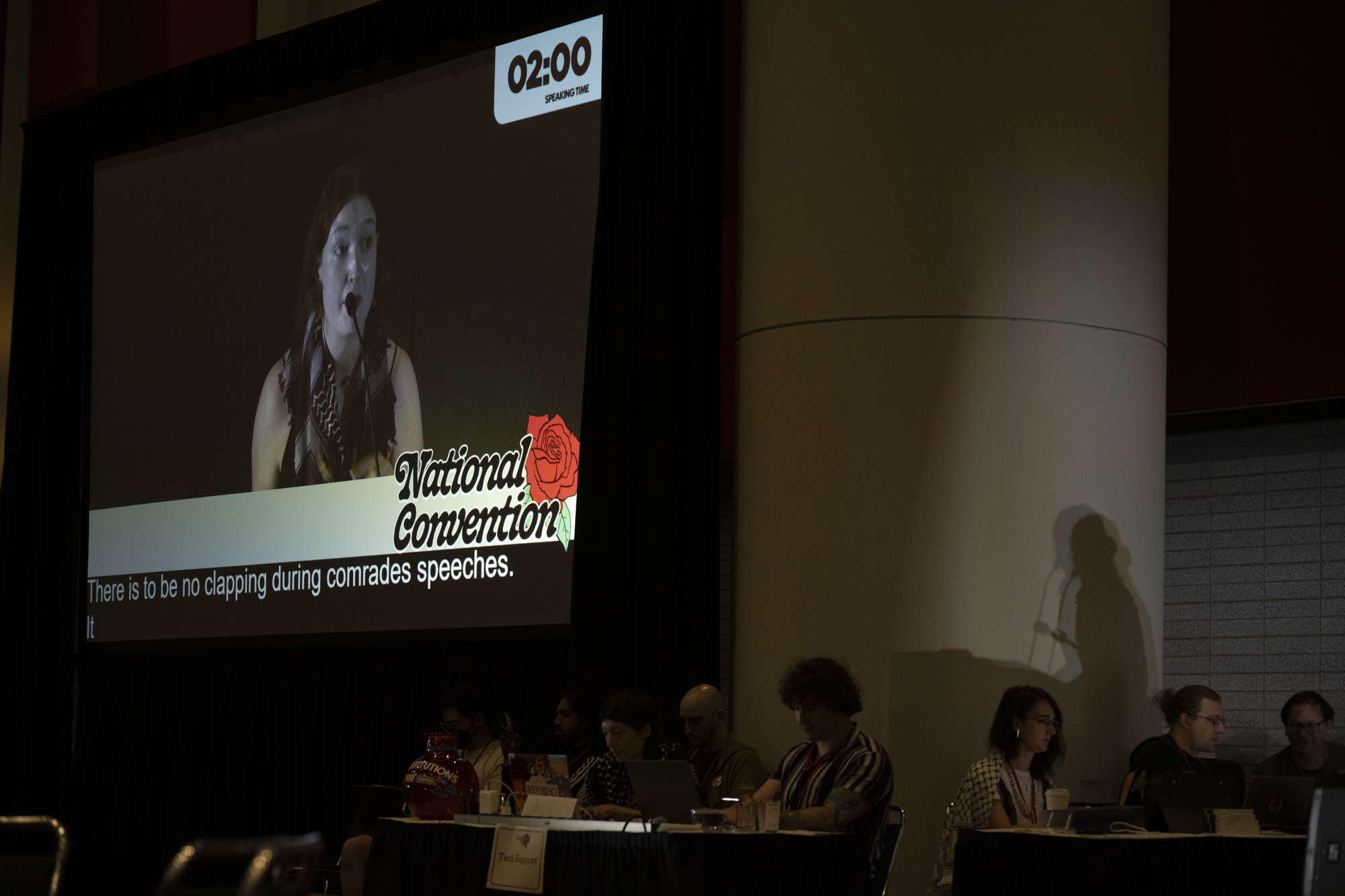 Silhouette of a woman speaking on a stage, seen on a large screen.