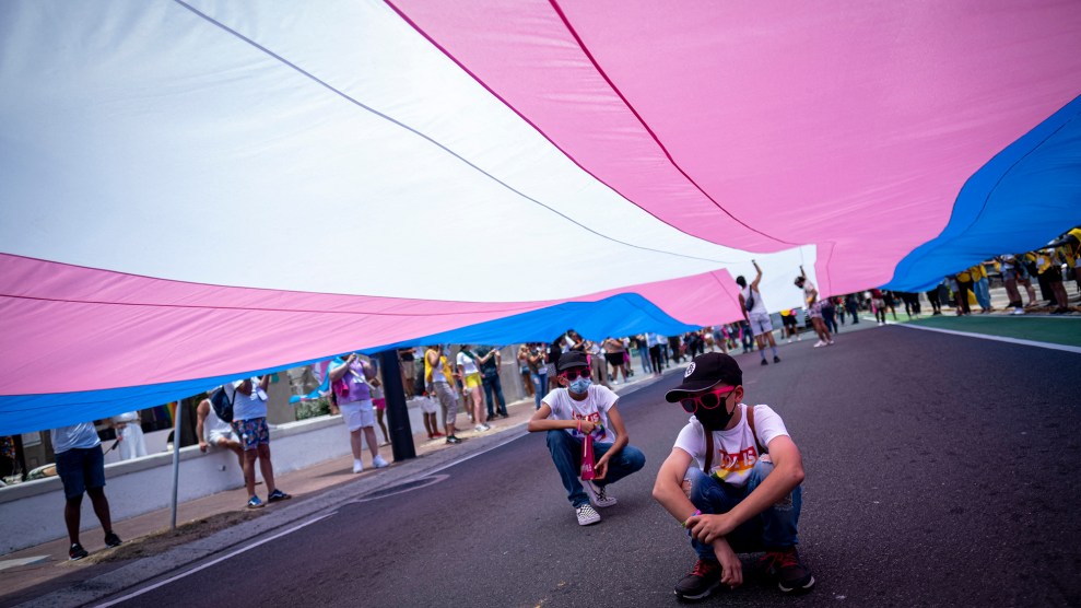 Two kids crouch under a large trans flag.