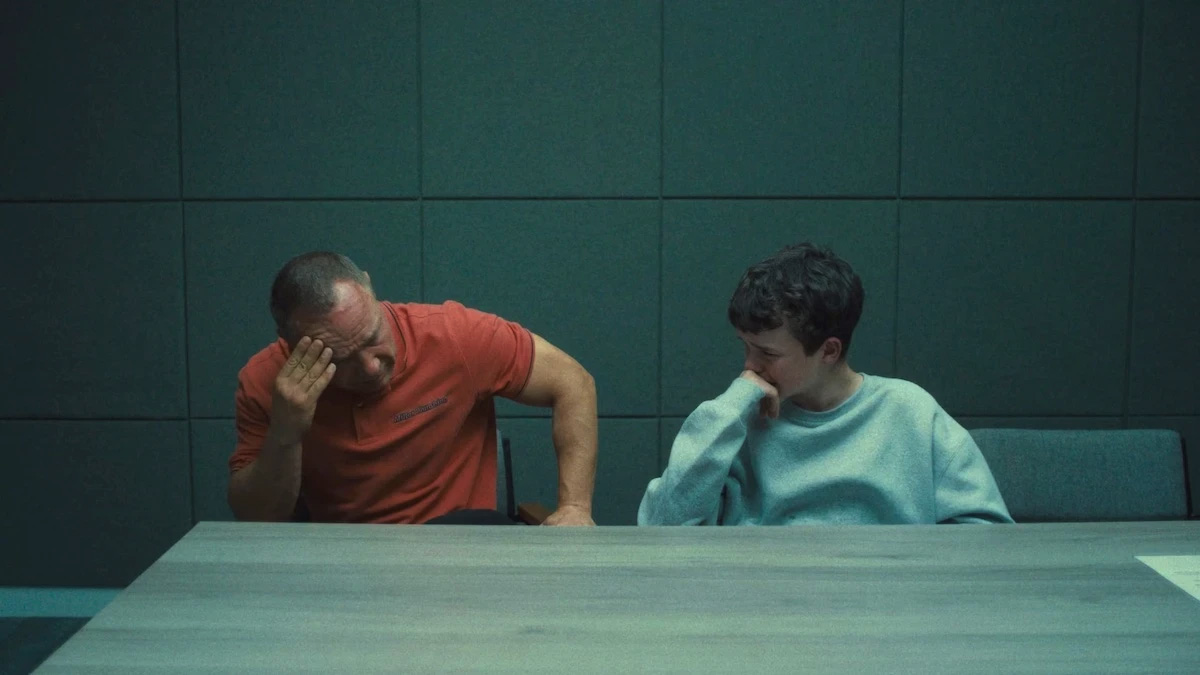 Young boy sitting next to his father at a table; still from TV show "Adolescence."