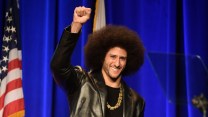 A biracial man with a large afro wearing a gold chain, black shirt, and a leather jacket, smiling and raising his right hand in a fist, standing in front of a blue background and an American flag