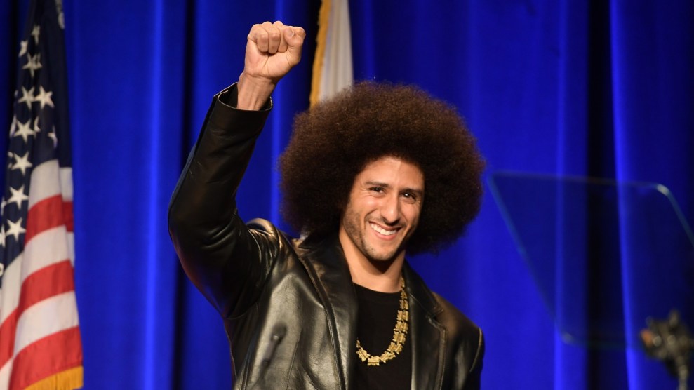 A biracial man with a large afro wearing a gold chain, black shirt, and a leather jacket, smiling and raising his right hand in a fist, standing in front of a blue background and an American flag