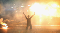 Seen from behind, an African American woman dressed in black is on her knees in front of a crosswalk in the street. In front of her is a bright, smoky explosion of orange, and beside her is an orange flare emitting smoke.