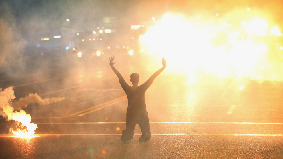 Seen from behind, an African American woman dressed in black is on her knees in front of a crosswalk in the street. In front of her is a bright, smoky explosion of orange, and beside her is an orange flare emitting smoke.