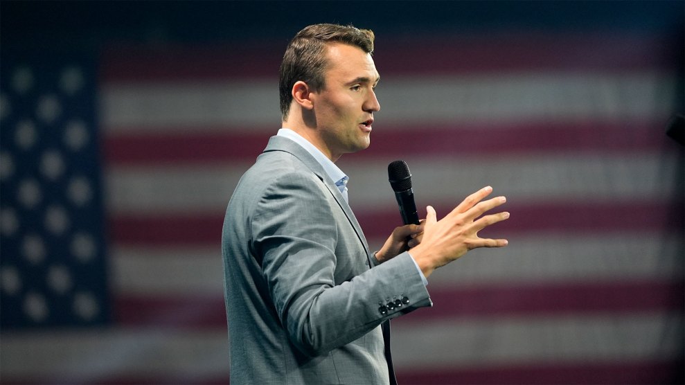 A man in a grey suit speaking into a microphone. A large American flag hangs in the background.