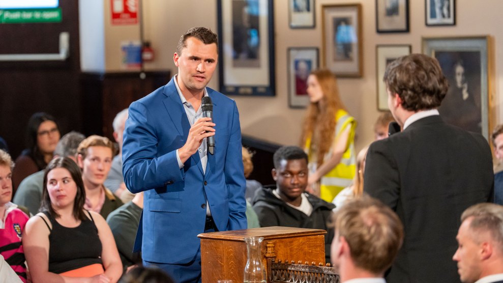 A man in a blue suit speaks into a microphone as a seated crowd of young people watches.