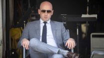 Stephen Miller sits in a gray suit and sunglasses, one leg crossed over the other