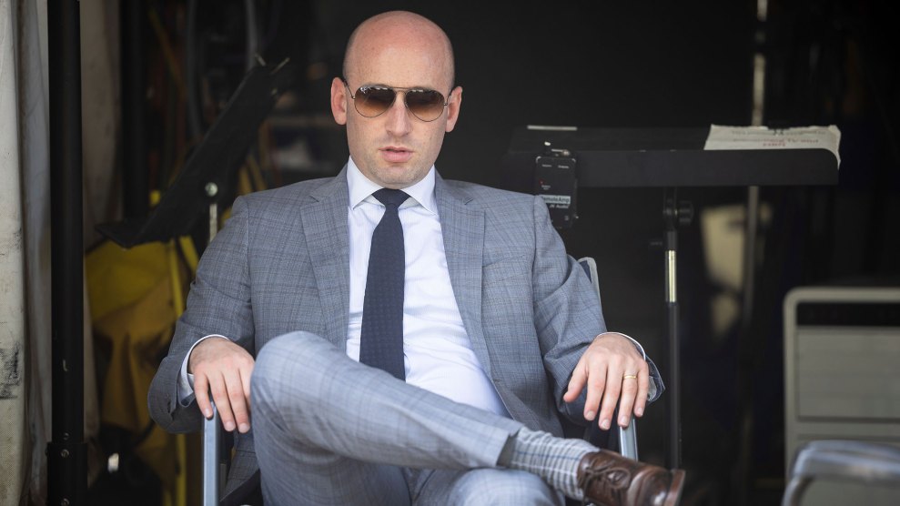 Stephen Miller sits in a gray suit and sunglasses, one leg crossed over the other