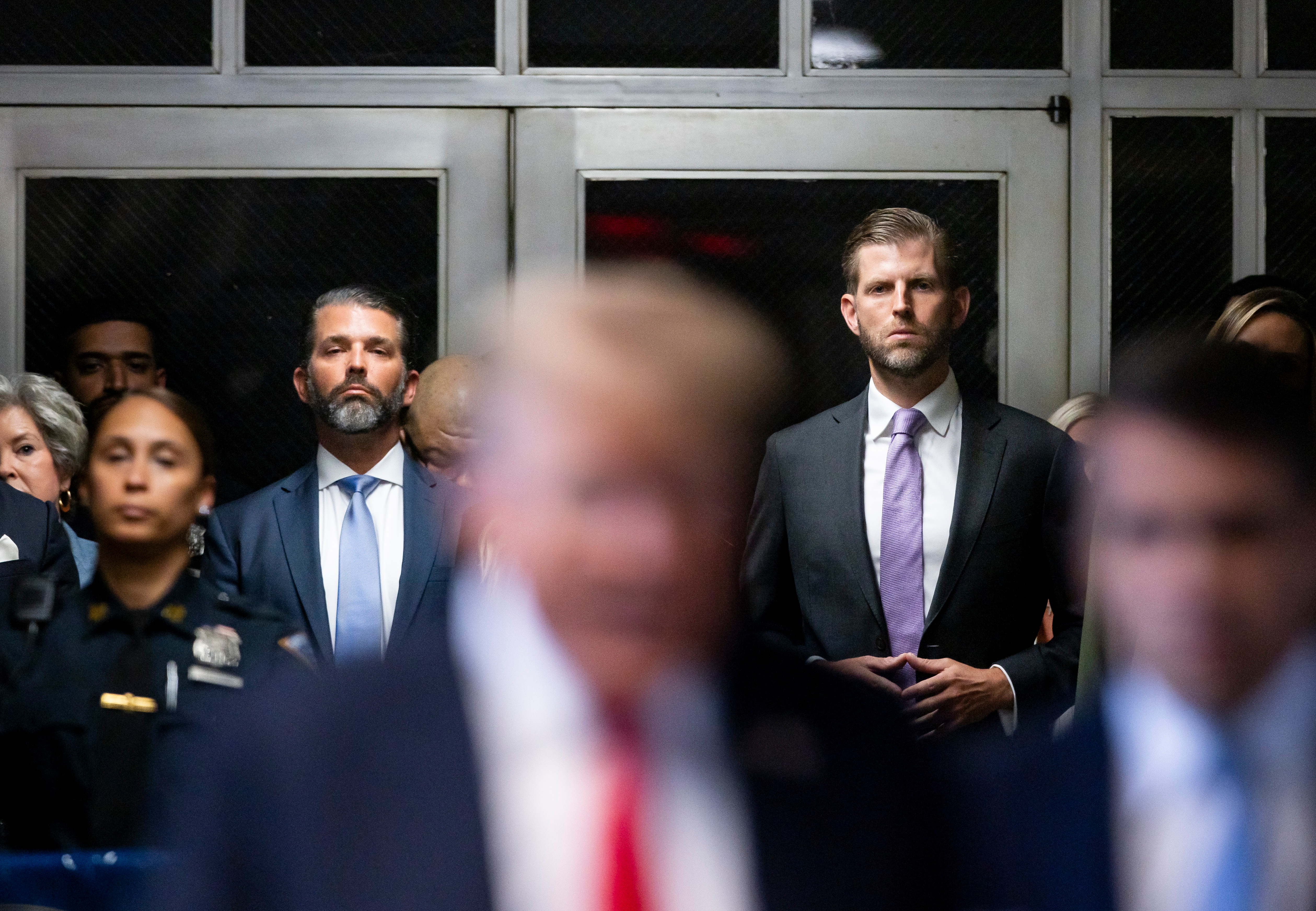 Donald Trump's two elder sons stand against two double doors. The two bearded men, seen in focus, look with rapt attention at their father, who is in the foreground but out of focus.