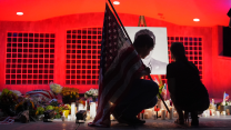 A man kneels while holding the American flag over his right shoulder amid flowers and candles in front of a black-and-white photo of Charlie Kirk. A woman kneels beside him.