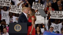 Donald Trump and Melania Trump share a moment on stage at a rally, surrounded by supporters holding 'Blacks for Trump 2020' signs.