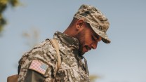 A picture of a bearded Black troop from the chest up, dressed in a camo hat and uniform with an American flag patch on his arm, facing profile to the left and staring at the ground