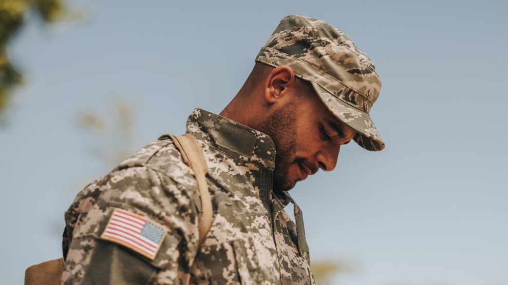 A picture of a bearded Black troop from the chest up, dressed in a camo hat and uniform with an American flag patch on his arm, facing profile to the left and staring at the ground