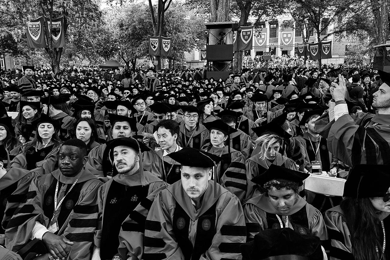 Black-and-white photo of a sea of robed Harvard grads facing the camera, seated in chairs on a lawn. Harvard veritas banners hang from trees in the background.