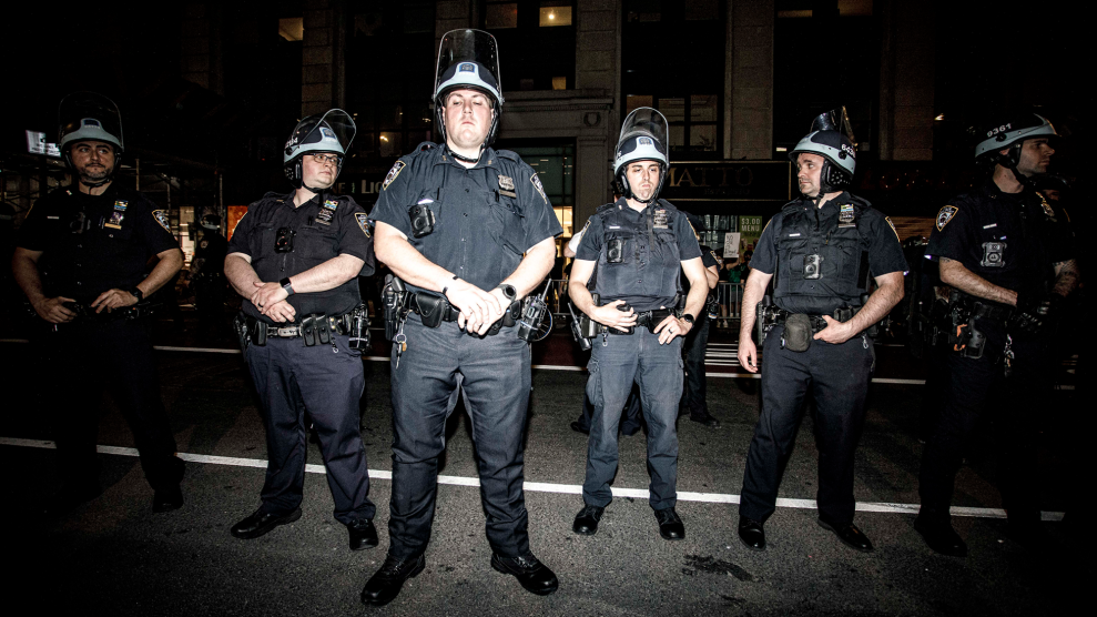 A row of six New York City police officers stand side by side at night under a bright flash of light. The officers are wearing helmets with face shields and are looking at each other.