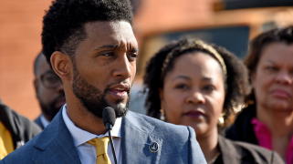 A young African American man with a closely trimmed beard, wearing a suit and tie, stands outdoors on a sunlit day behind a microphone as he looks into the distance. Several African American men and women stand behind him.