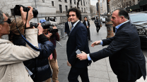A clean-shaven man with dark curly hair, wearing a dark suit and tie, looks into the camera as photographers and reporters surround him on a New York City sidewalk. A man in a suit behind him holds up his hands, as if attempting to clear a path.
