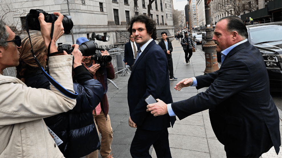 A clean-shaven man with dark curly hair, wearing a dark suit and tie, looks into the camera as photographers and reporters surround him on a New York City sidewalk. A man in a suit behind him holds up his hands, as if attempting to clear a path.