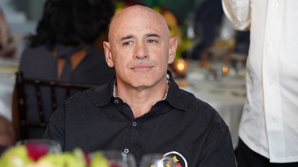 A ruddy, bald-headed white man with large ears sits at a table with wine glasses, dressed in a black Pittsburgh Steelers button-up shirt.