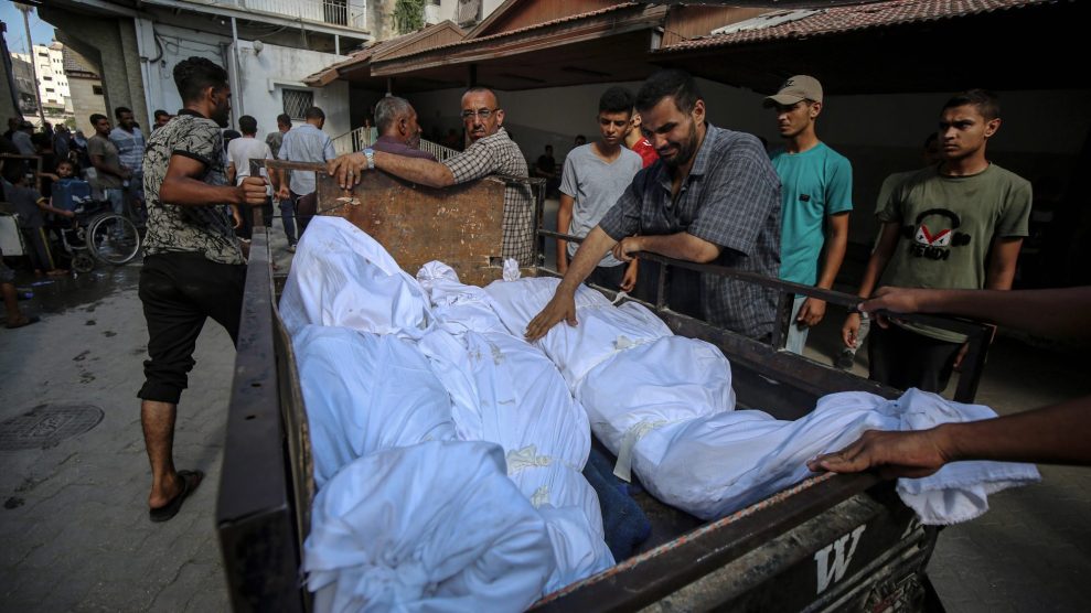 A group of men surround a truck with bodies in the background wrapped in white cloths.