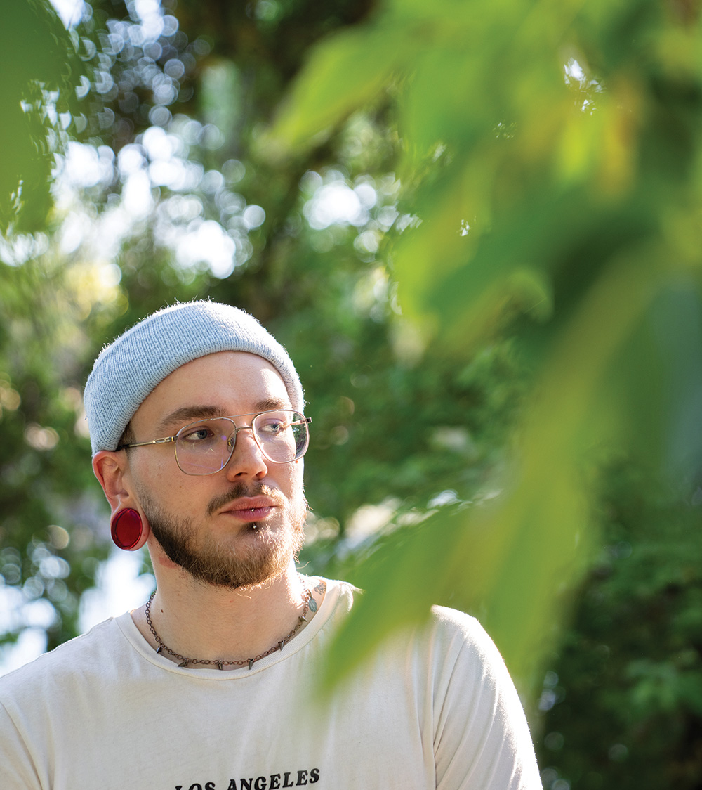 Portrait of a young Caucasian man wearing a pale blue beanie, wire-rim glasses, and white T-shirt. He has a short, dark beard and a lip piercing, and a large red gauge is visible in his left earlobe.