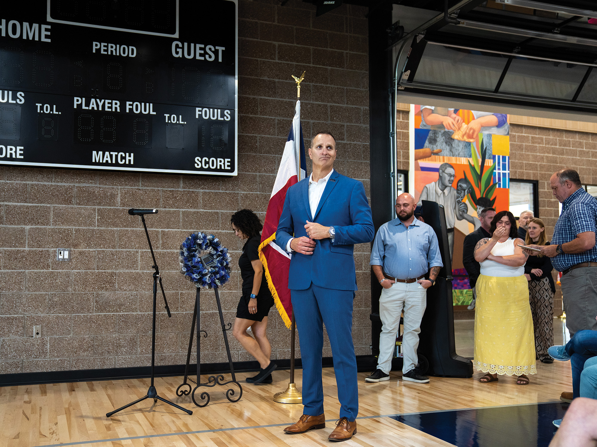 A tall Caucasian man with a buzz cut, wearing a blue suit, stands in a gymnaisum. His facial expression is a half-smirk.