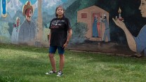Dee Maes-Sandoval, dressed in shorts and a T-shirt, stands in front of a painted mural. The mural depicts a woman in a white shawl holding a red rose, a man wearing a beige cowboy hat, and a house where a man hands a baby to a woman standing in the doorway.