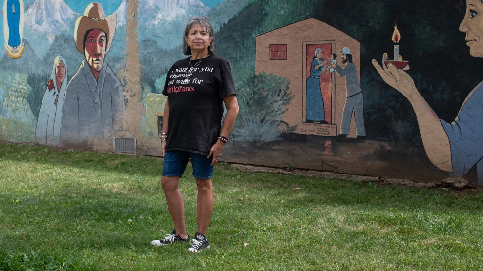Dee Maes-Sandoval, dressed in shorts and a T-shirt, stands in front of a painted mural. The mural depicts a woman in a white shawl holding a red rose, a man wearing a beige cowboy hat, and a house where a man hands a baby to a woman standing in the doorway.