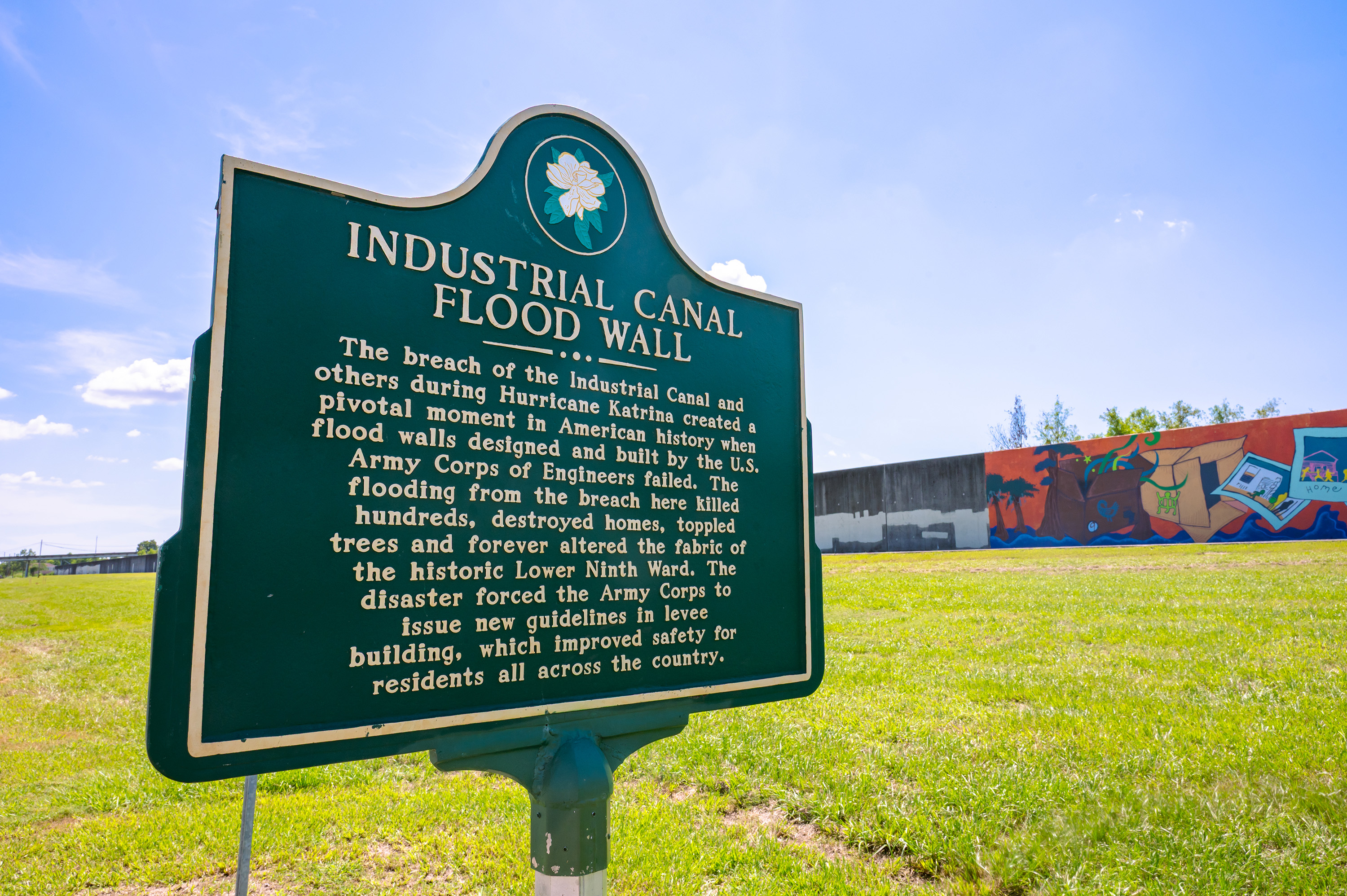 A green sign stands in a field near a floodwall with a brightly painted mural. The sign reads, "Industrial Canal Flood Wall," and summarizes the devastation that occurred when Hurricane Katrina breached the walls at the location.