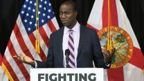 A man in a suit and striped tie, Dr. Joseph Ladapo, stands at a podium with the word “FIGHTING” printed on the front. He gestures with both hands, palms up, and has an uncertain, questioning expression on his face. Behind him are the U.S. flag and the Florida state flag.