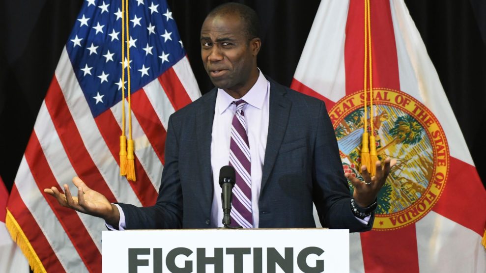 A man in a suit and striped tie, Dr. Joseph Ladapo, stands at a podium with the word “FIGHTING” printed on the front. He gestures with both hands, palms up, and has an uncertain, questioning expression on his face. Behind him are the U.S. flag and the Florida state flag.