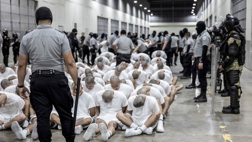 Men sit huddled on a concrete floor of a prison, surrounded by guards. The men all wear white T-shirts, shorts, socks, and clogs and have their heads shaved.
