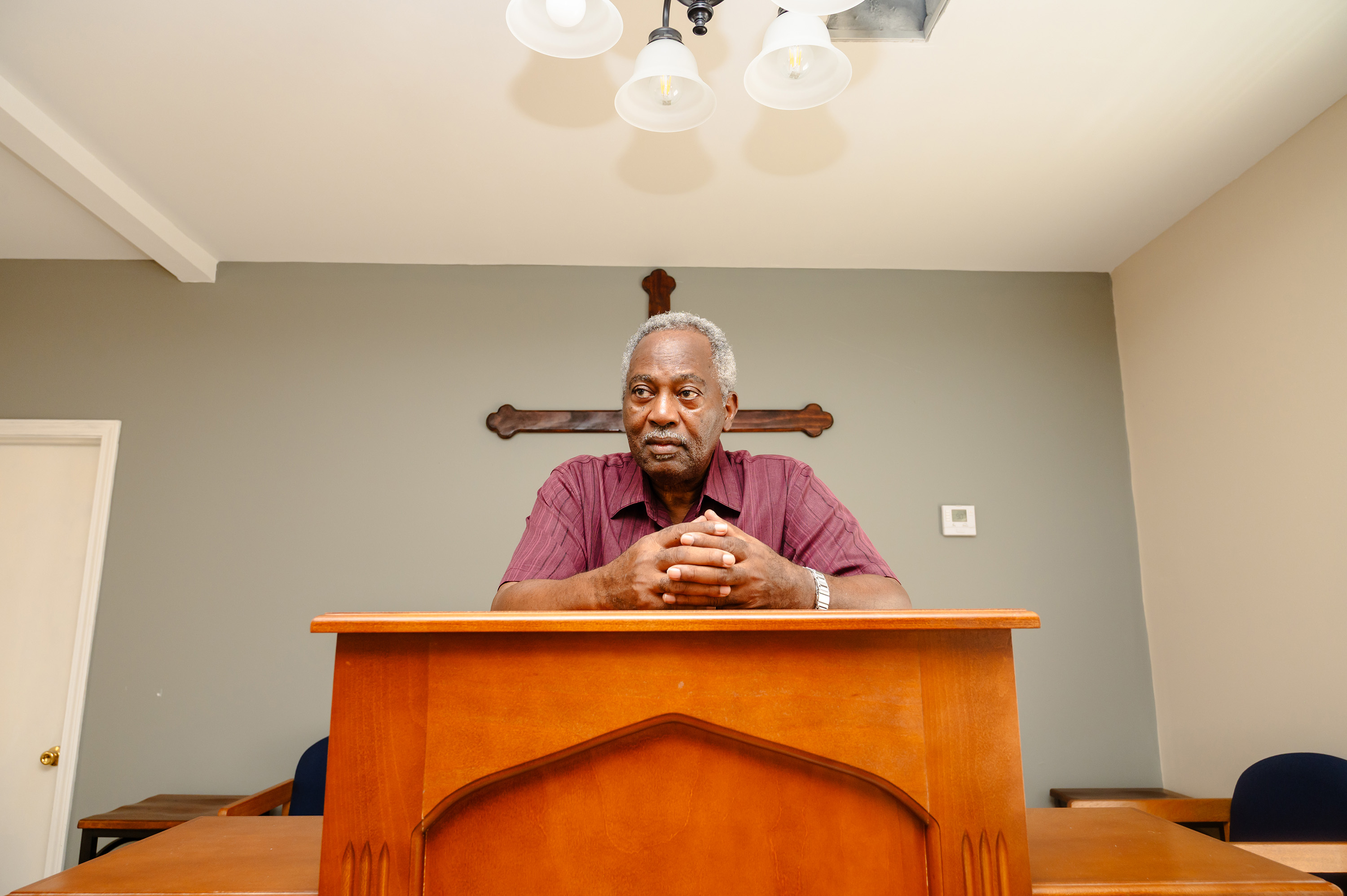 An older African American man leans over pulpit. A wooden cross hangs on the wall behind him.
