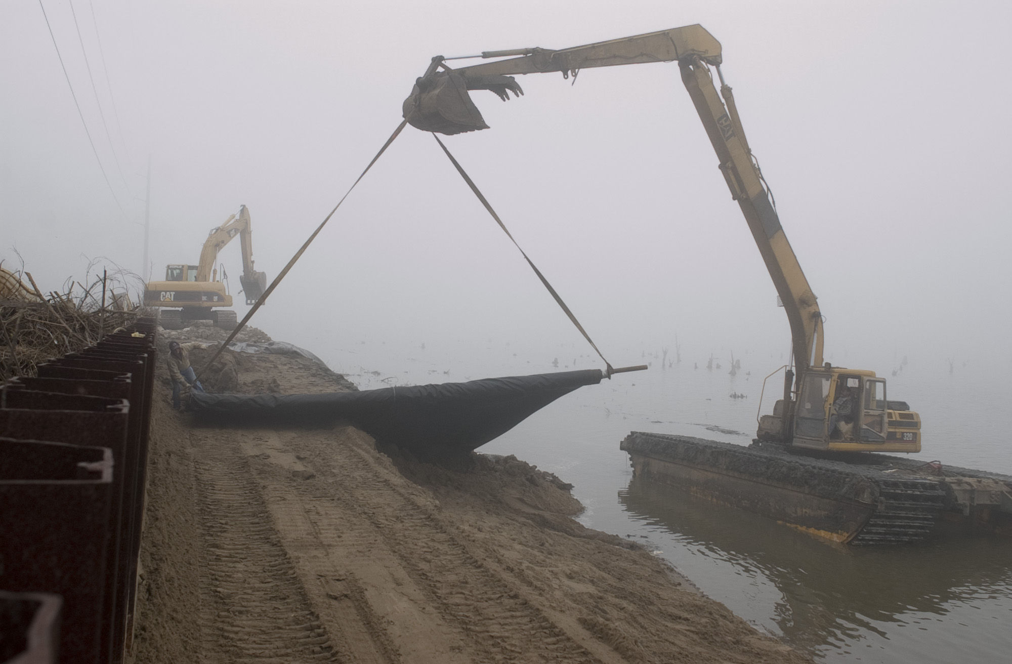 A crane sitting in murky water lowers a large tarp over the edge of a sandy embankment.