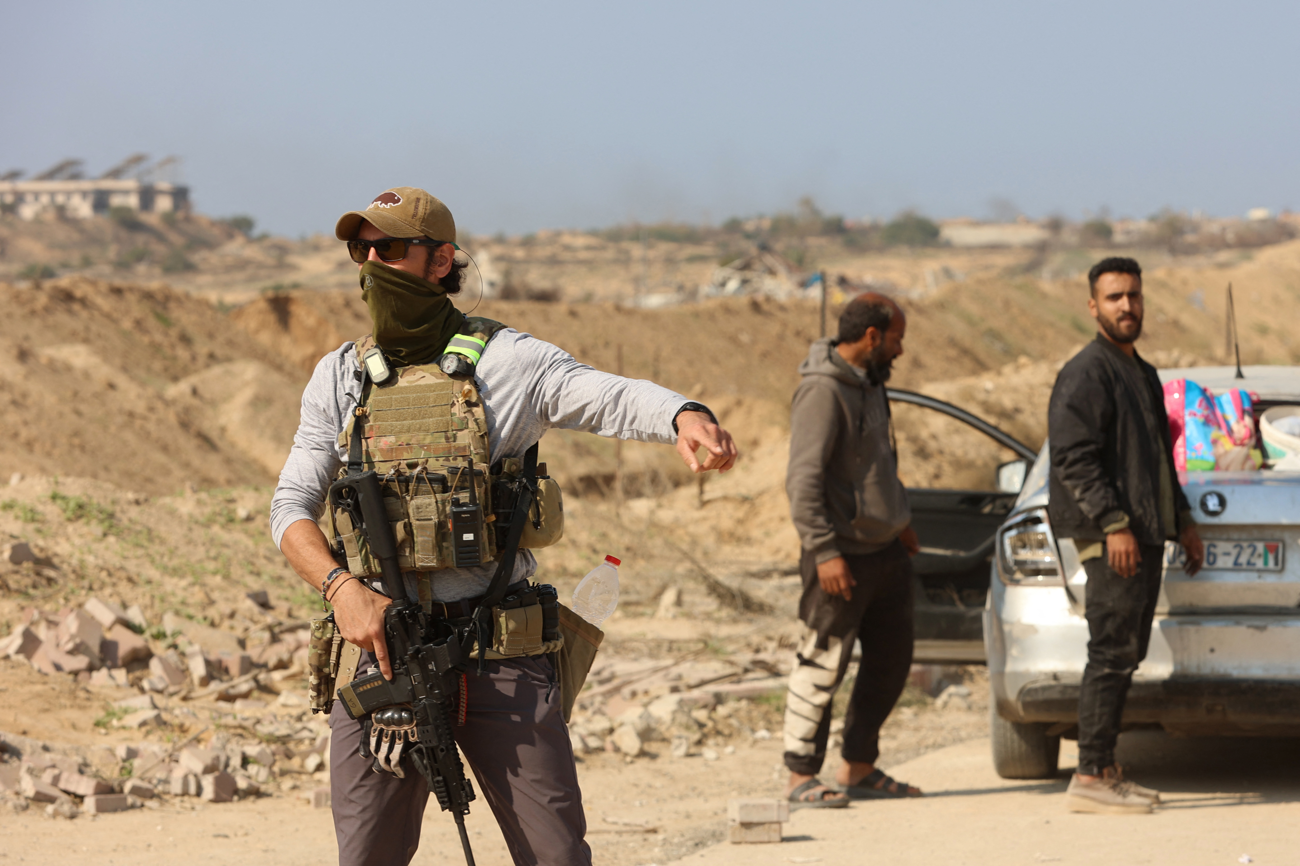 An armed man in a tactical vest, sunglasses, baseball cap, and face mask points toward a road, directing Palestinians where to go. 