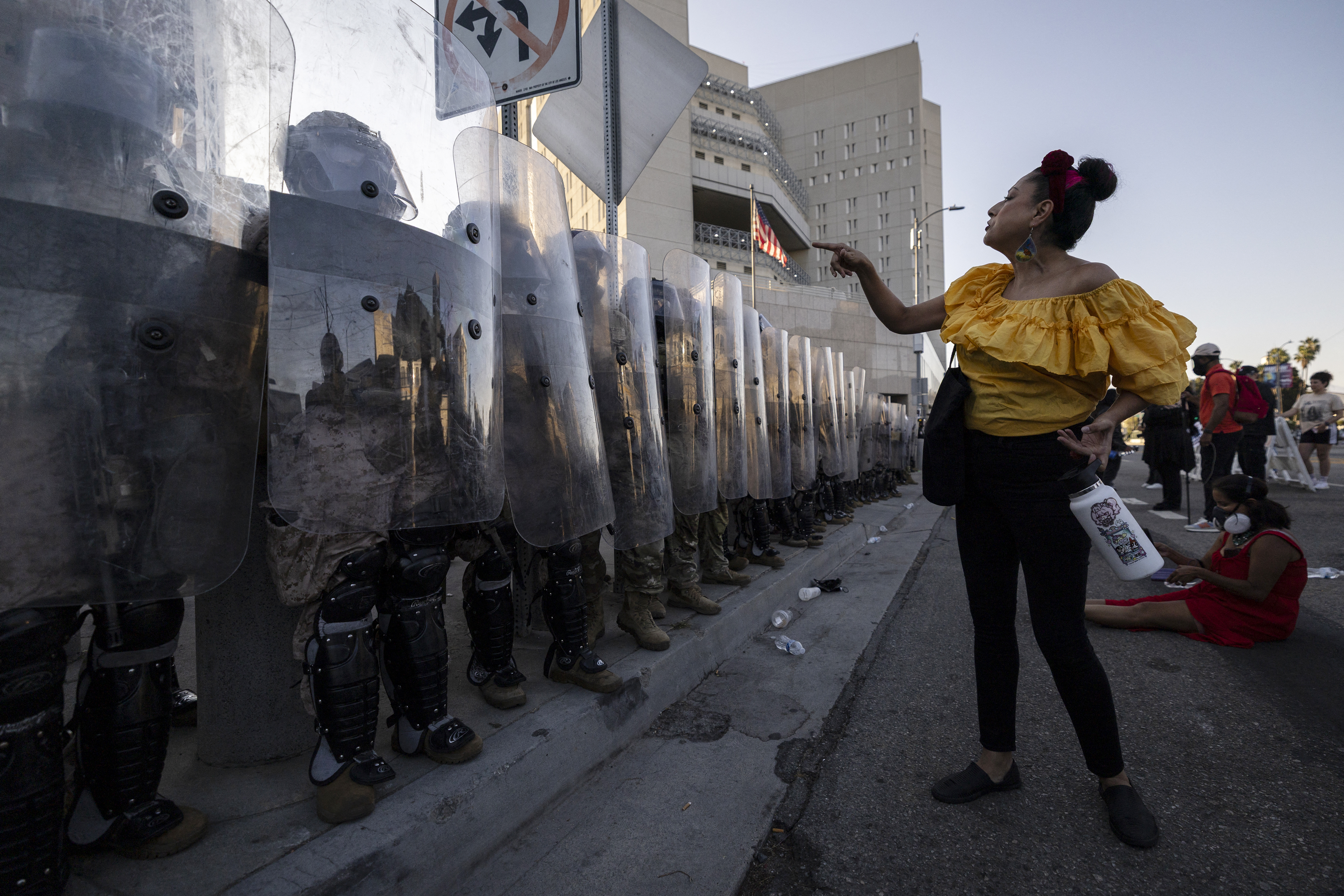 A woman in a ruffled yellow shirt confronts in front of a line of troops dressed in riot gear and holding up clear shields. 