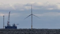 An offshore wind mill stands in the ocean with a construction ship to its left.