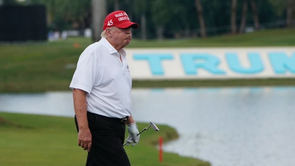 Donald Trump wearing a red hat, white polo, black slacks, and holding a golf putter. He is walking across a green. Behind him, a flower bed spells the word "TRUMP."