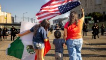 A family of three stands in front of armed, black-clothed riot police. We see the family from behind, the mother hoisting an upside down US flag, while a daughter holds one from Mexico.