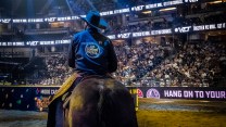 Seen from behind in a rodeo arena, a man sits astride a horse in a dark cowboy hat and blue buton-up shirt that reads, "Protected by US Border Patrol."