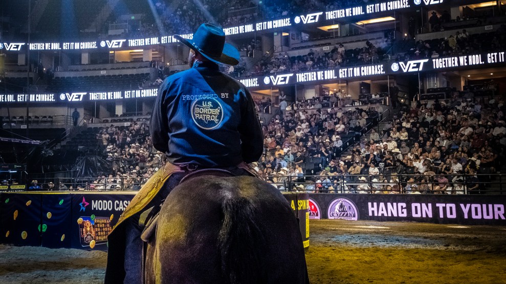 Seen from behind in a rodeo arena, a man sits astride a horse in a dark cowboy hat and blue buton-up shirt that reads, "Protected by US Border Patrol."