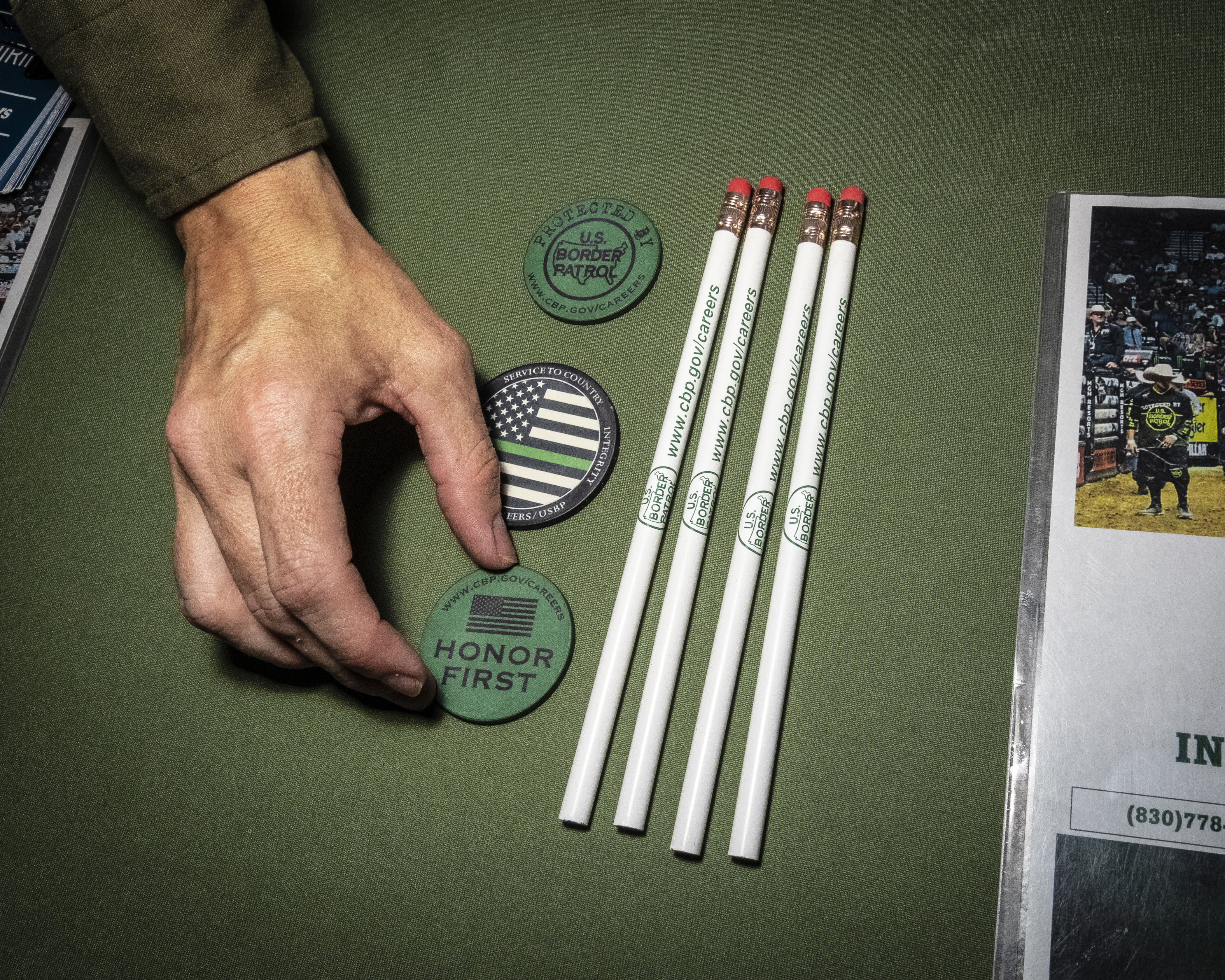 A hand reaches for a green plastic token that reads, "Honor First," next to four white CBP-branded pencils on a table.