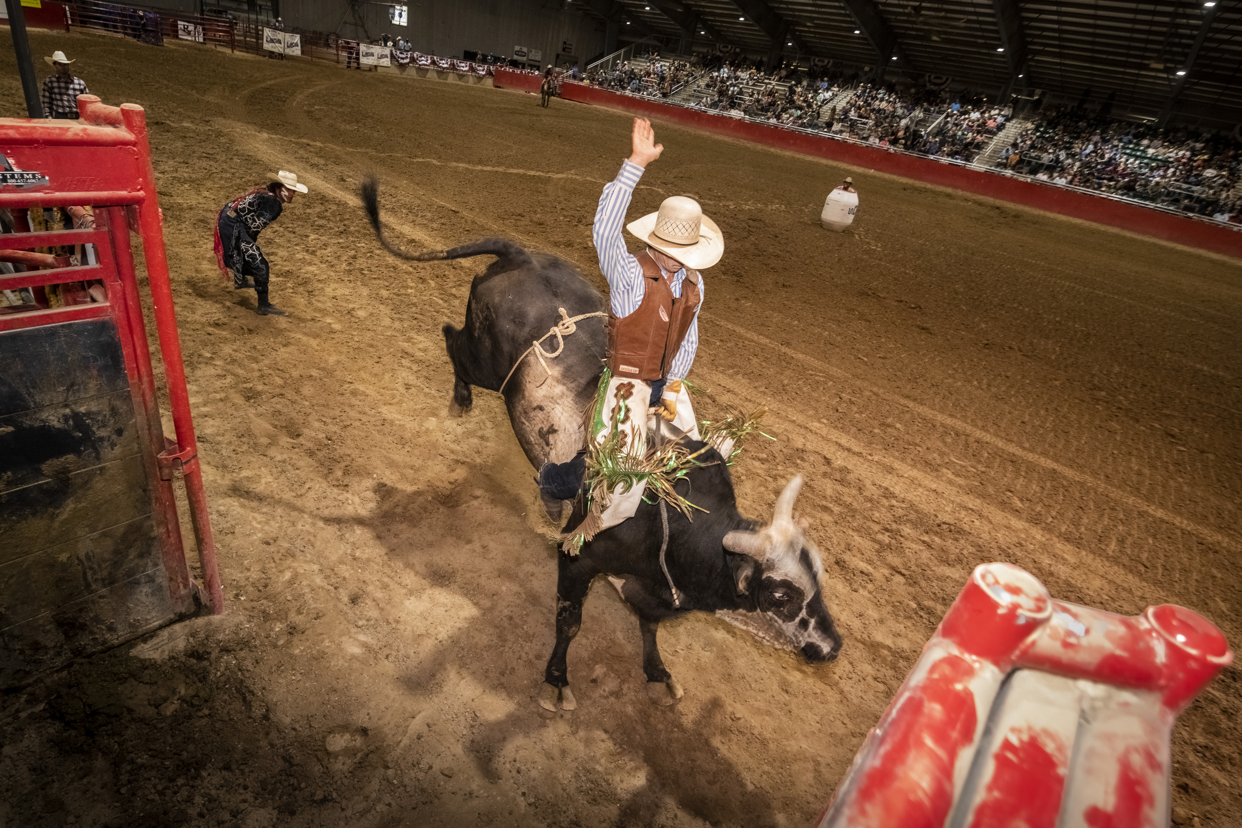 A man, whose face is hidden by light beige cowboy hat, rides a bull in a rodeo. His right hand is raised high in the air.
