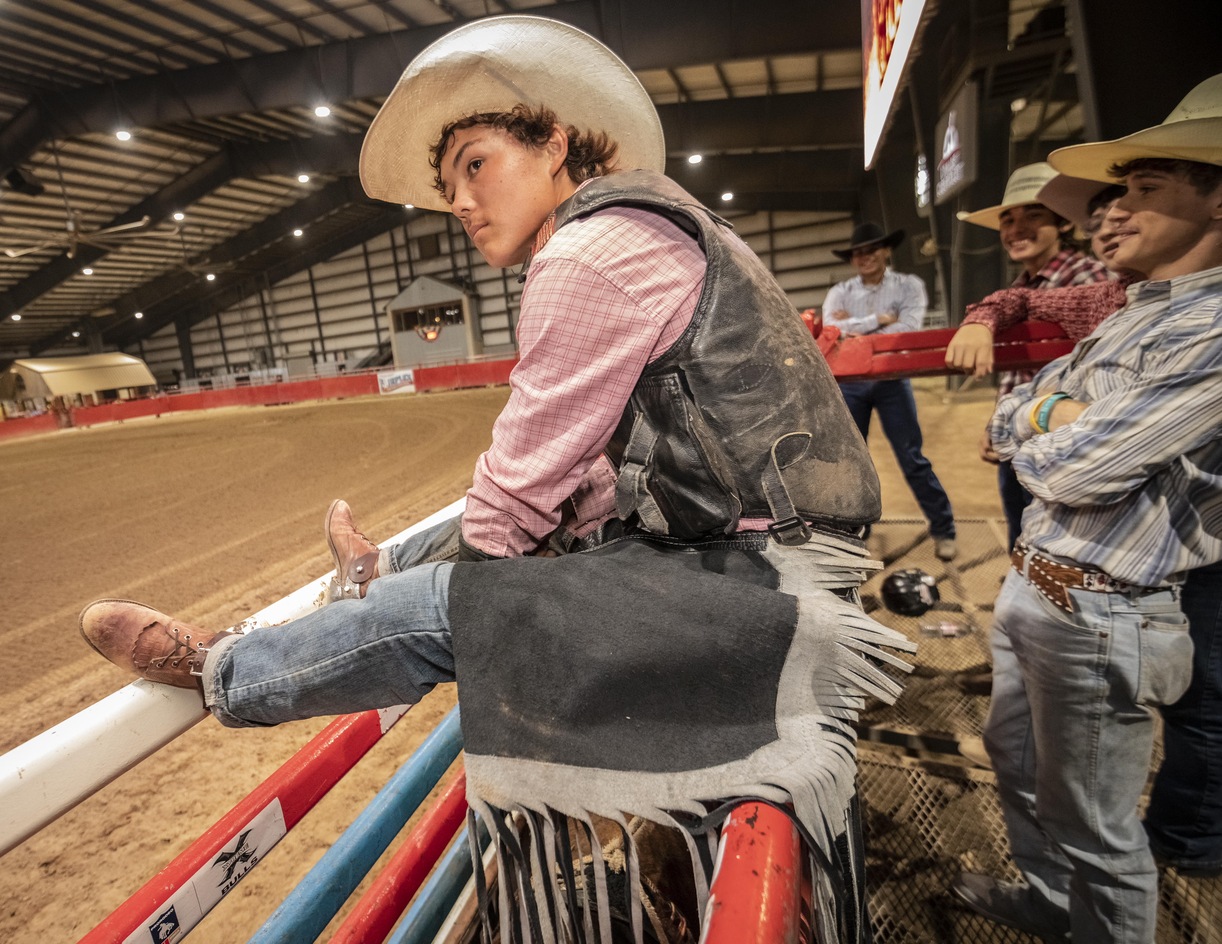 A young man in a white cowboy hat, black leather vest, pink plaid shirt, black chaps, and jeans sits on a metal gate at a rodeo.