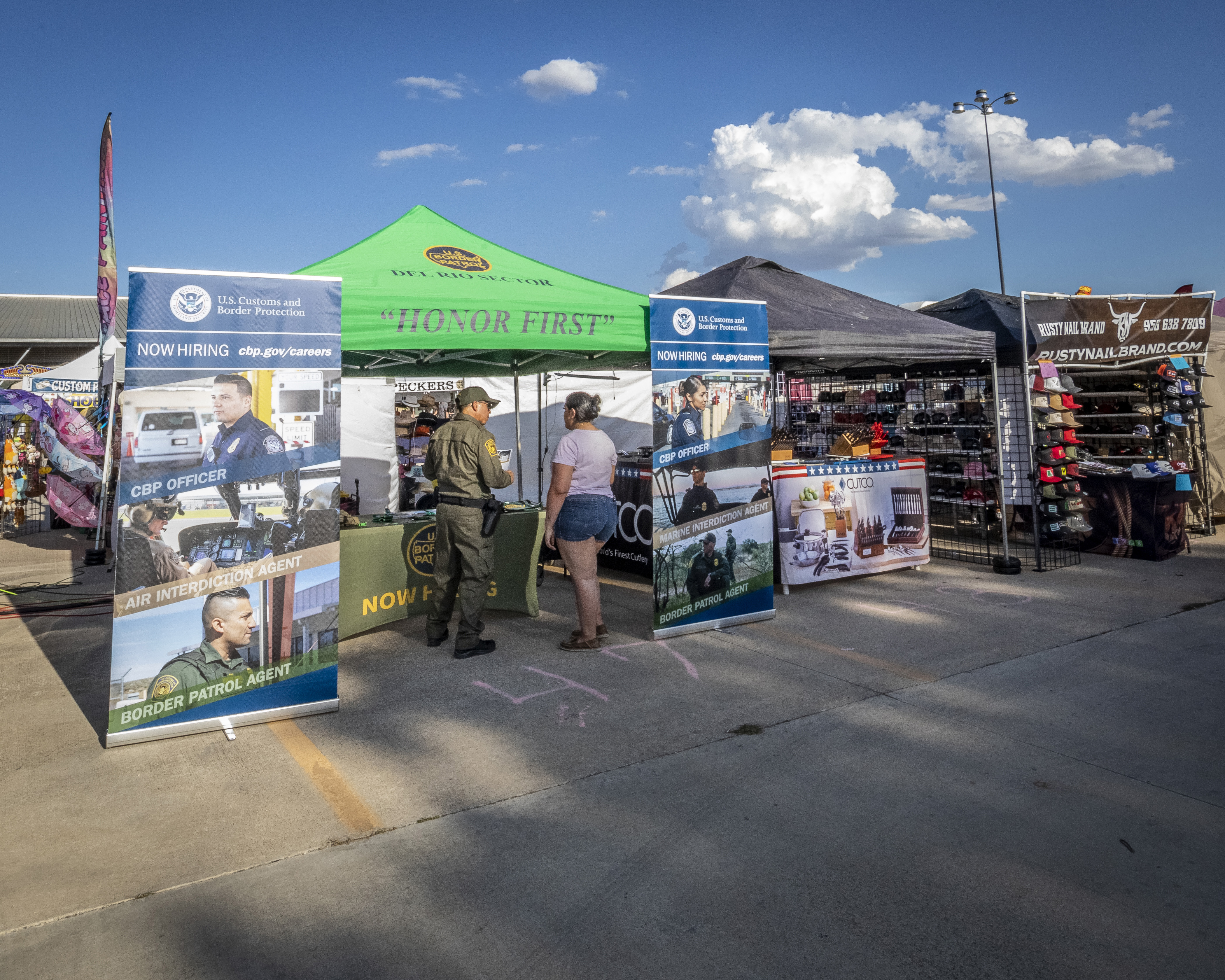 A man in an olive green Border Patrol uniform talks to a woman under a green pop-up canopy that reads, "Honor First." Large advertisements for CBP flank the canopy.