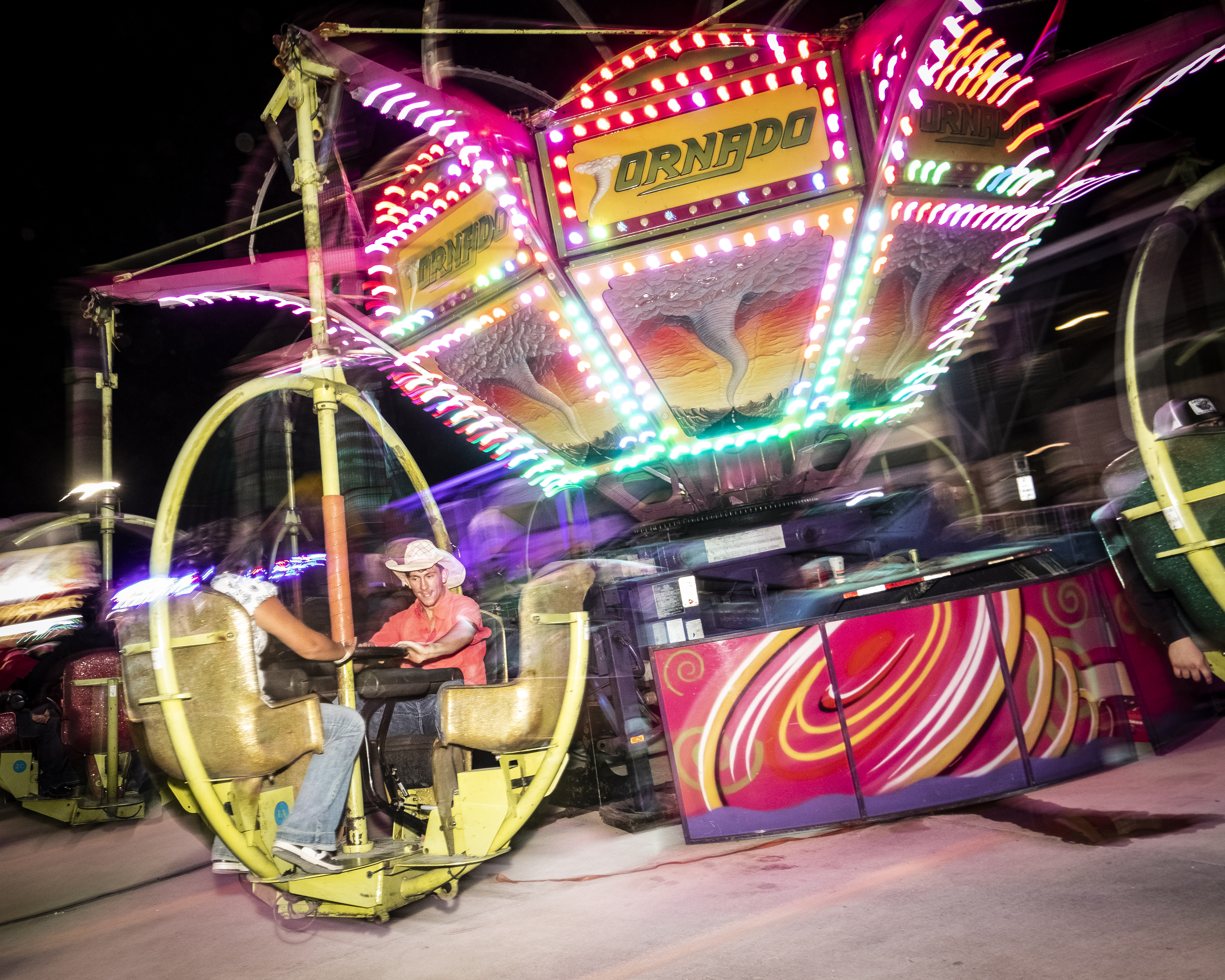 Two men on a brightly colored carnival ride called "Tornado" at night.