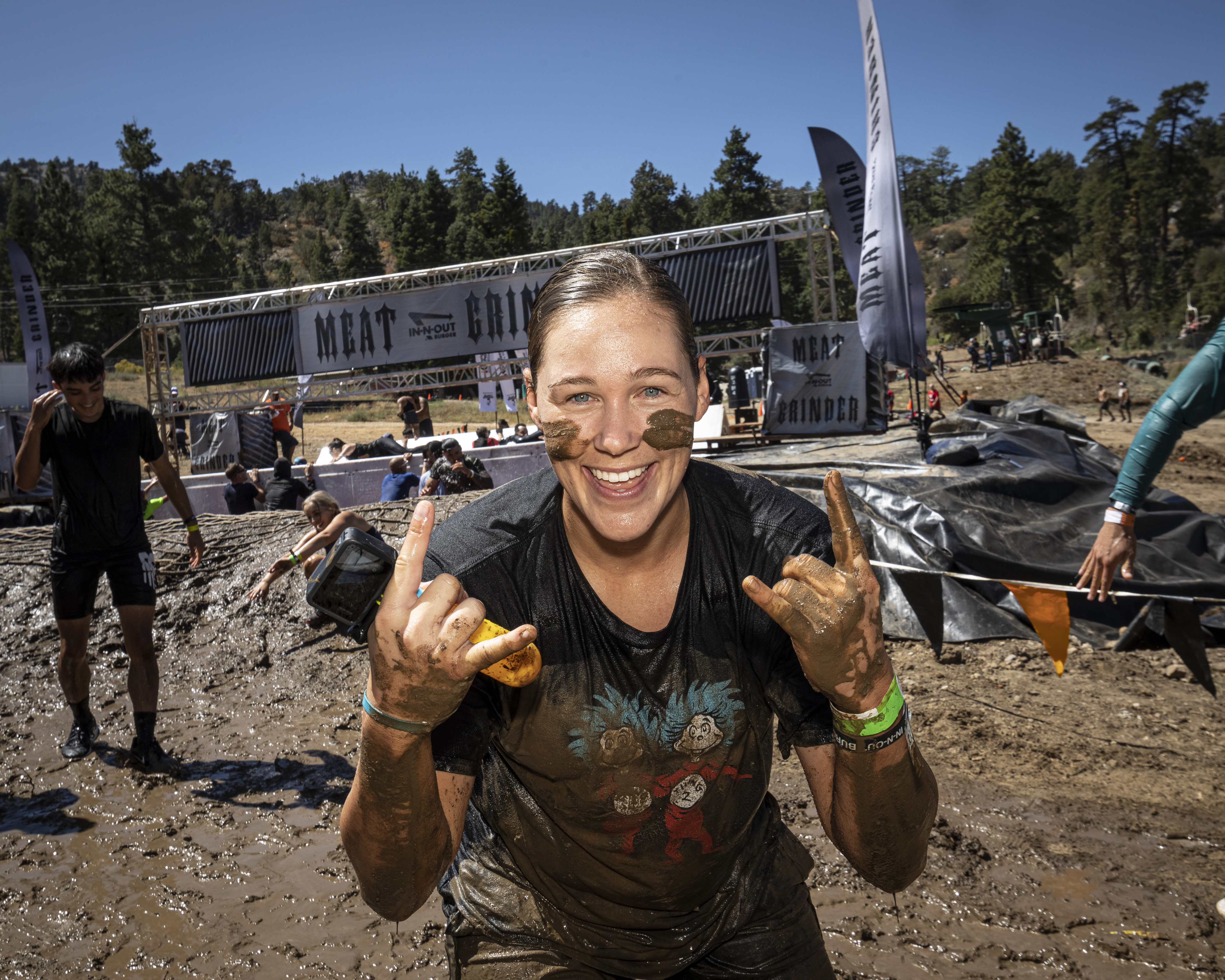 A woman covered in mud smiles and makes "rock on" gestures with both hands.