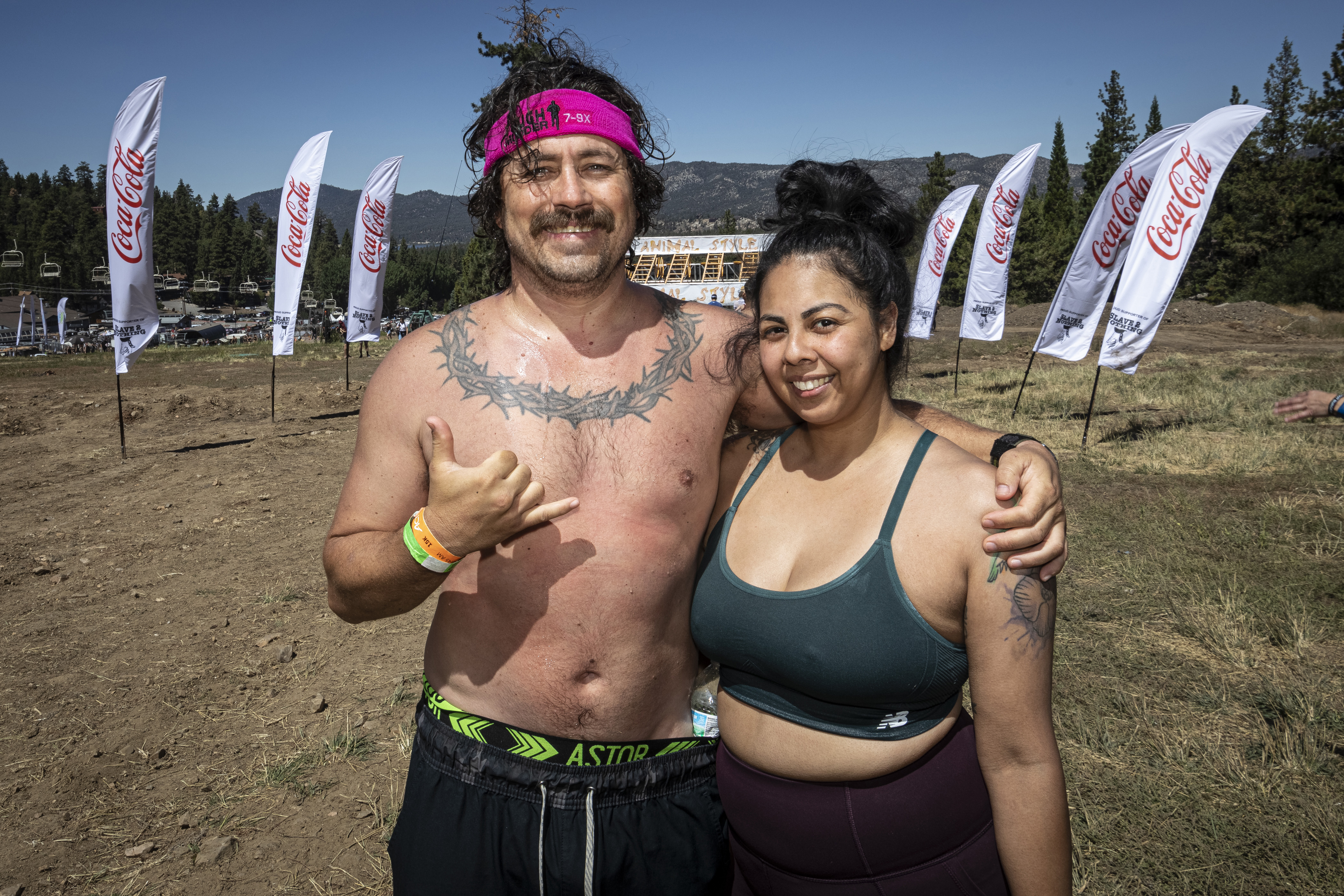 A shirtless man stands with his arm around a woman in a cropped sports tank top. The man makes a shaka hand gesture with his right hand.