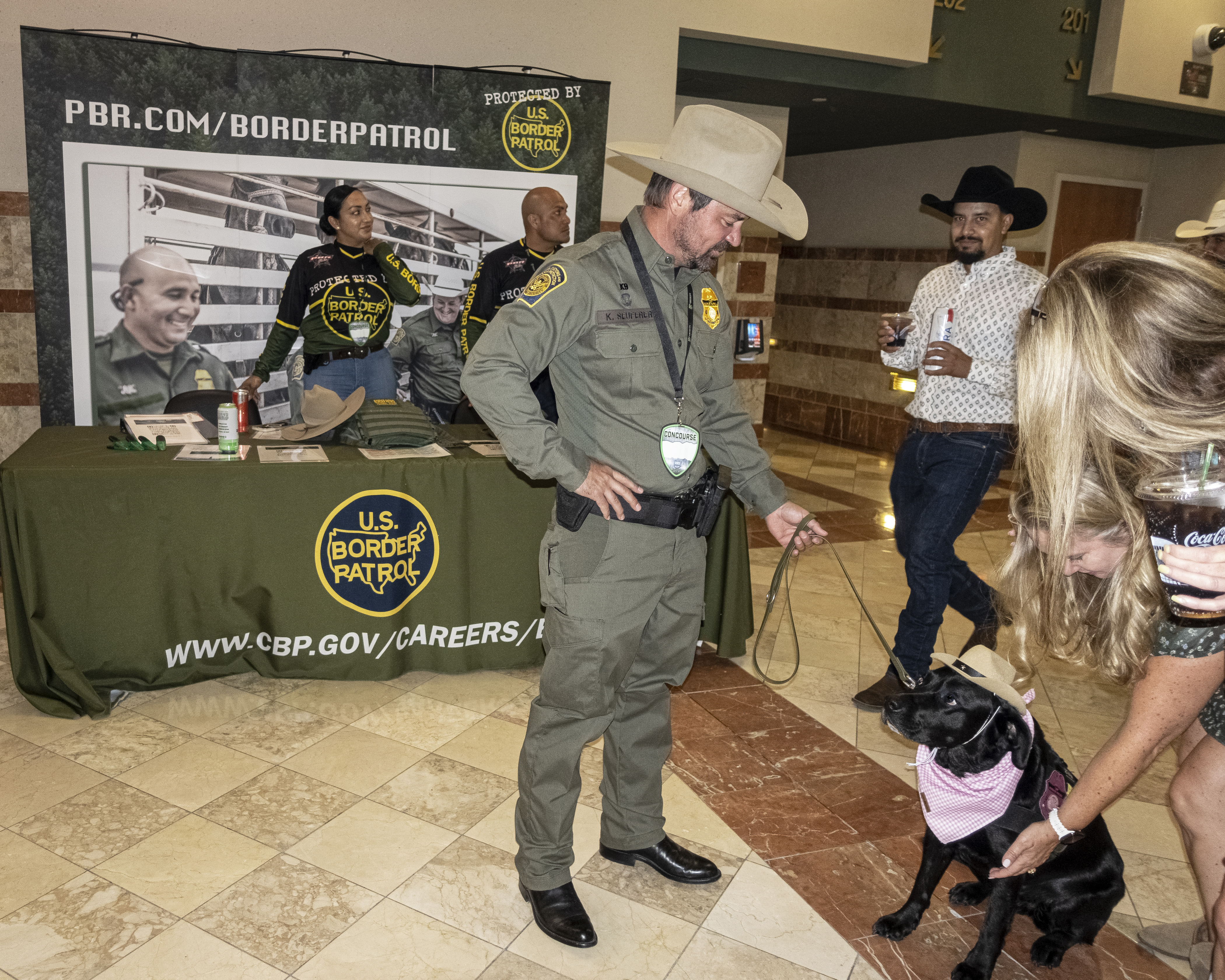 A uniformed Border Patrol agent holds the leash of a black dog, which wears a tiny cowboy hat and gingham bandanna. A woman with blond hair bends down to pet the dog.