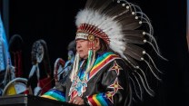 A man wears a headdress and a rainbow-adorned outfit while speaking at a lecturn.