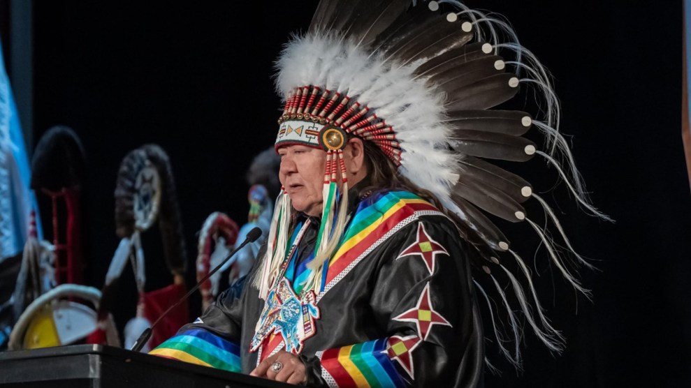 A man wears a headdress and a rainbow-adorned outfit while speaking at a lecturn.